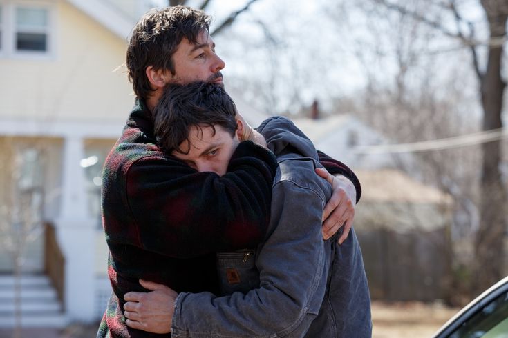 two men, likely a father and son, are hugging each other tightly in a show of deep emotion and support. the older man, with a beard, is comforting the younger man, who appears to be upset or in distress. the setting is outdoors on a cold winter day, with trees in the background and a house and car visible in the distance. the father's plaid jacket and the son's blue jacket suggest a sense of casual warmth and togetherness.  their body language conveys a powerful message of love, care, and empathy during a difficult moment.