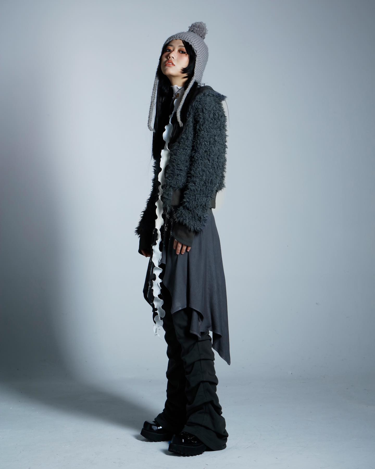 a fulllength studio shot of a young woman showcasing a fashionable outfit. she wears a grey fluffy jacket, a dark grey asymmetrical skirt, black gathered pants, black platform boots, and a grey beanie with long ear flaps. a white ruffled accessory hangs down from her waist. the overall style is dark, edgy, and alternative, with a unique layered aesthetic.  the background is a plain light grey. the model is posed threequarter view with her body slightly turned.