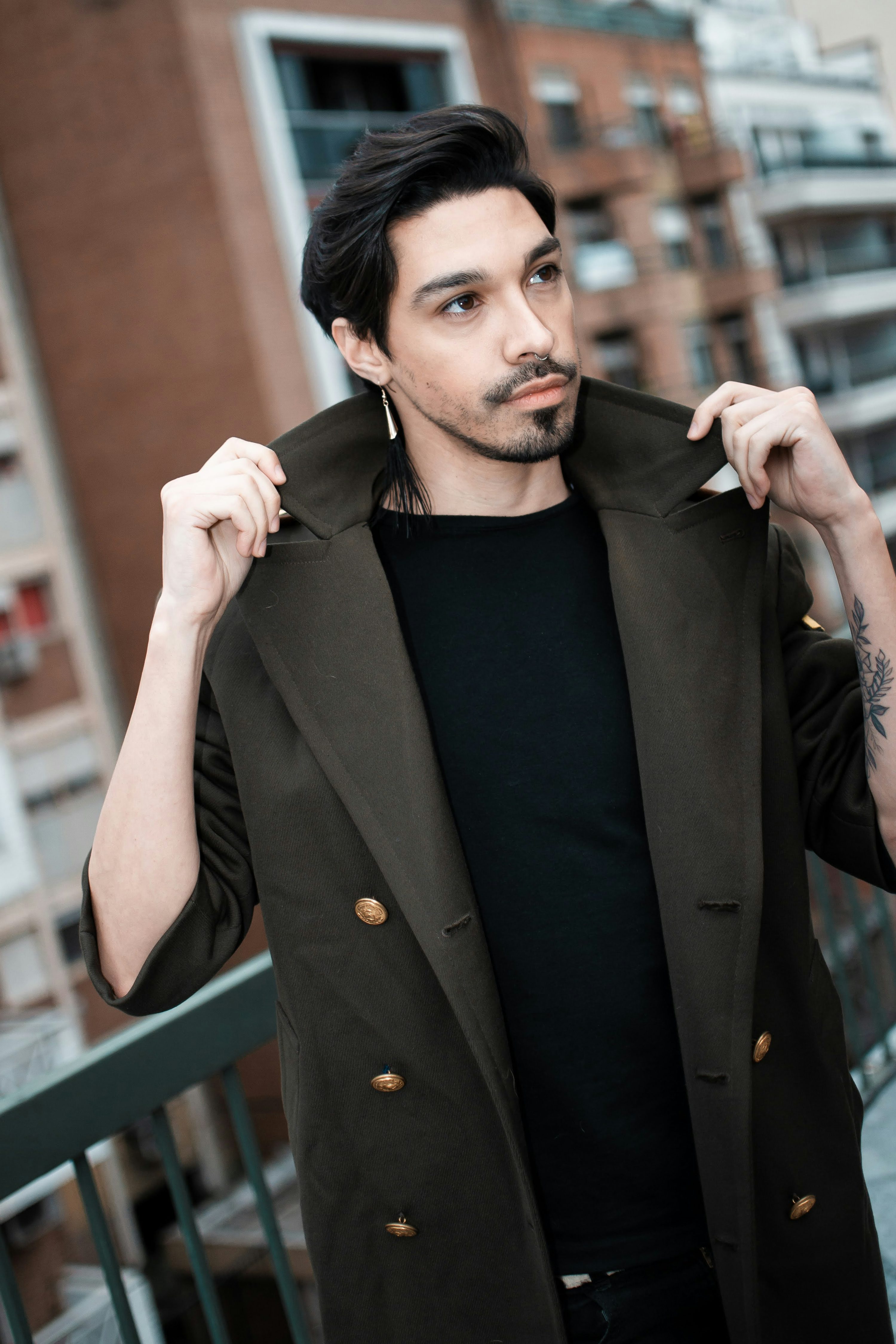a stylish man with long dark hair is standing outdoors, wearing a doublebreasted olive green coat over a black shirt. he's posing confidently, adjusting his coat collar with one hand. the background is a blurred city scene with brick buildings. he's wearing long earrings. the overall impression is one of urban, fashionable cool.