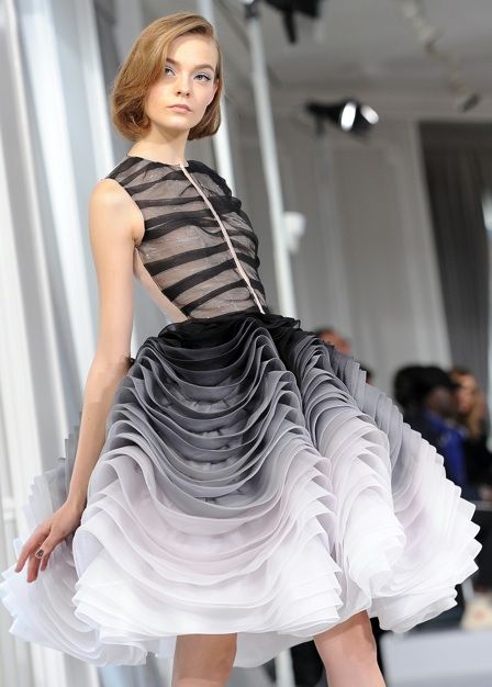 a fashion model walks the runway in a stunning black and white ombre layered dress. the dress is made of a sheer and flowing fabric that creates a cascade of ruffles and gathers. the model's hair is pulled back in a simple style, allowing the dress to be the focus of attention. the overall effect is elegant and chic, with a touch of drama and sophistication.