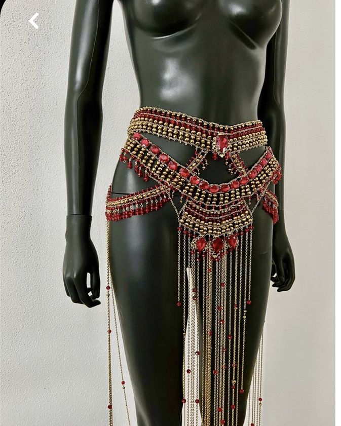 a black mannequin is wearing a gold and red beaded and jeweled waist belt. the belt has multiple layers of beads and jewels, with chains hanging down from the bottom layer.