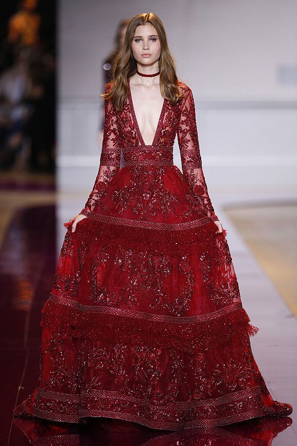 a woman in a red gown with intricate embroidery and sequins walks down the runway. she has a plunging neckline and long sleeves. the dress is layered and flows gracefully. the model also has a choker necklace.