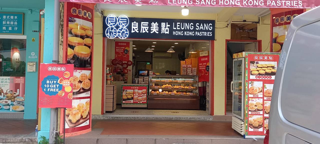 this image shows the storefront of a hong kong bakery called leung sang hong kong pastries. there are several pastries and desserts on display inside the shop and on the signs outside. the bakery has a traditional look with bright red and gold accents, and the sign above the door is in chinese characters that translate to "leung sang." the image captures a moment of everyday life in hong kong, where local bakeries are known for their delicious and traditional pastries.
