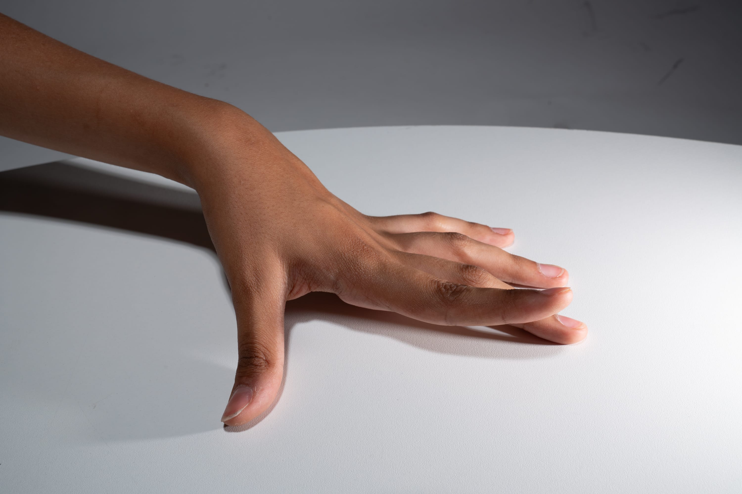a hand with brown skin is gently placed on a white table with its fingers spread out. the lighting is soft, creating a subtle shadow under the hand.