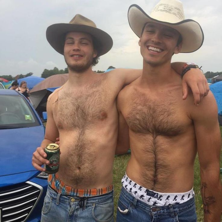 the image shows two young, shirtless men embracing outdoors. both men have significant body hair on their chests and are wearing cowboy hats. one man is holding a beer can. they appear to be at a festival or outdoor event, with tents and a blue car visible in the background. the overall vibe is relaxed and carefree. they are smiling and appear to be good friends.