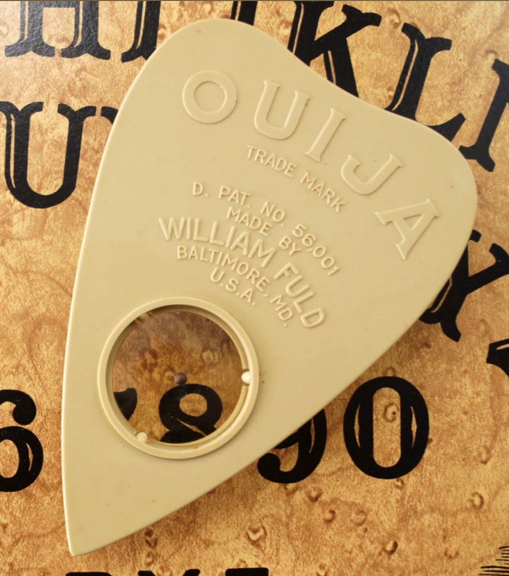 a closeup image of an antique ouija board planchette. the planchette is a heart shape with a clear window, featuring a brown dot in the middle, and the words "ouija", "trade mark",  "d. pat. no. 56,001", "made by",  "william fuld", "baltimore, md.", and "u.s.a." are printed on it.  the planchette is sitting on a wooden ouija board with a number of letters and numbers visible in the background. the image suggests a sense of mystery, and the occult.