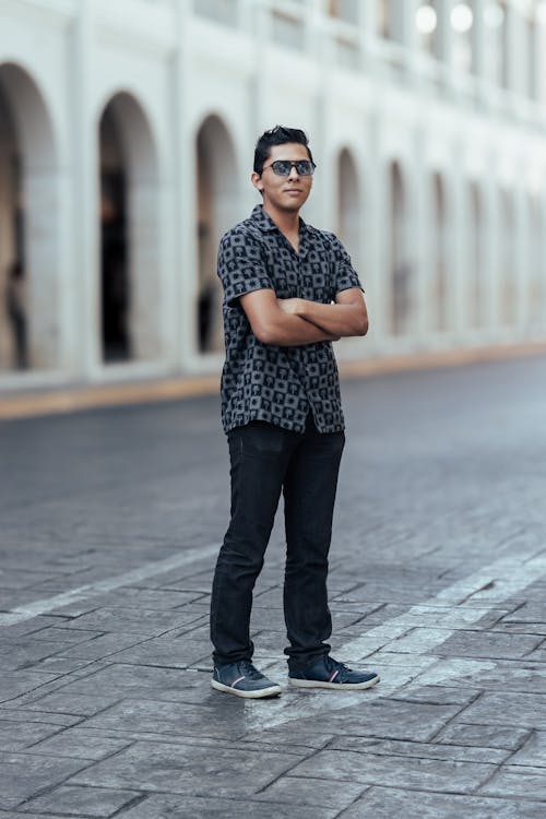 a young man with dark hair and sunglasses stands confidently on a city street with his arms crossed. he is wearing a patterned gray shortsleeved shirt, dark jeans, and dark shoes. the background is blurred but shows a row of classical style buildings with arches. the overall impression is one of stylish casual wear in an urban setting.