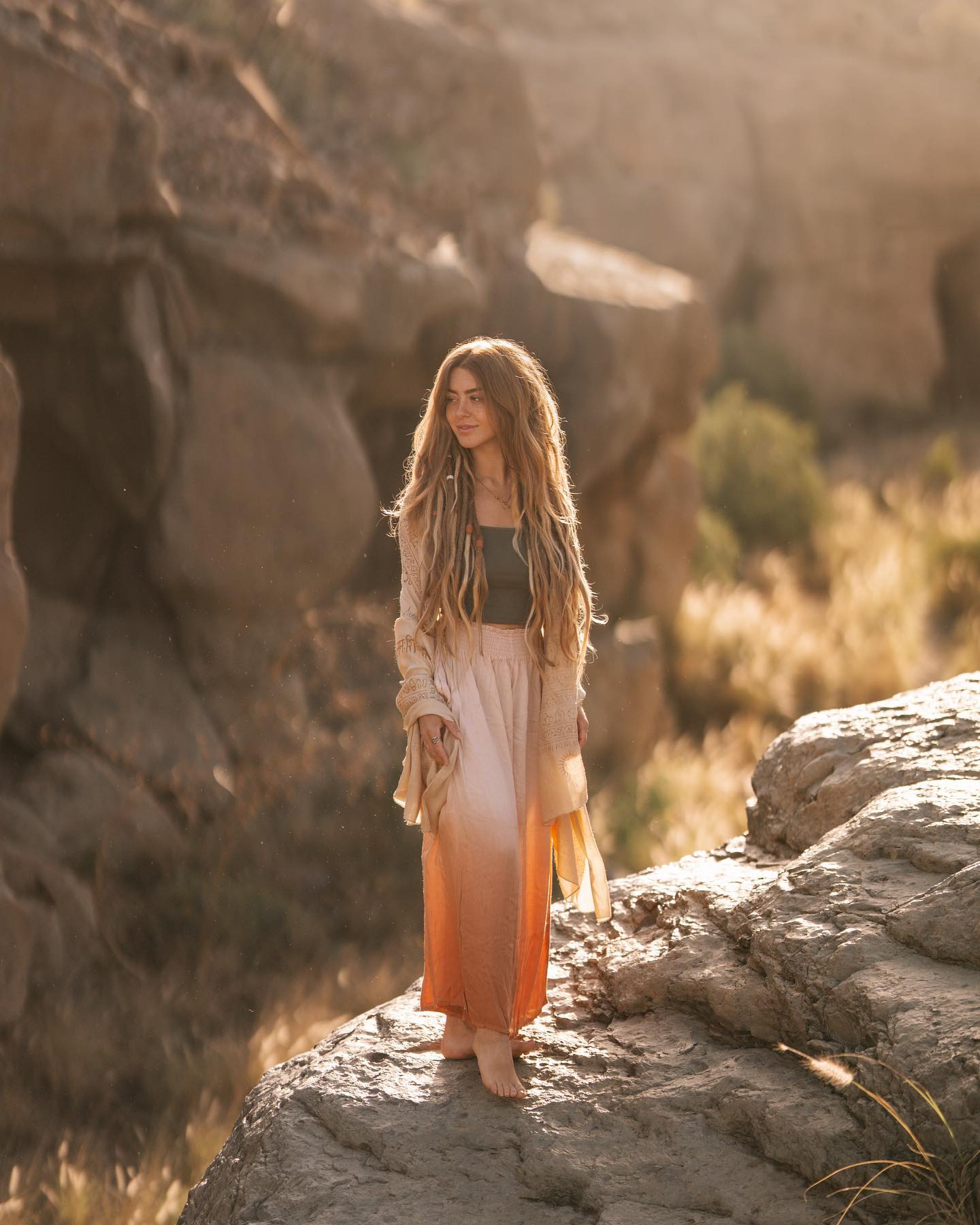 a young woman with long dreadlocks stands on a rocky outcrop in a desert landscape.  she's wearing a peach ombre maxi skirt and a light beige cardigan. the setting sun casts a warm golden light on her and the surrounding scenery. the overall mood is serene and peaceful.