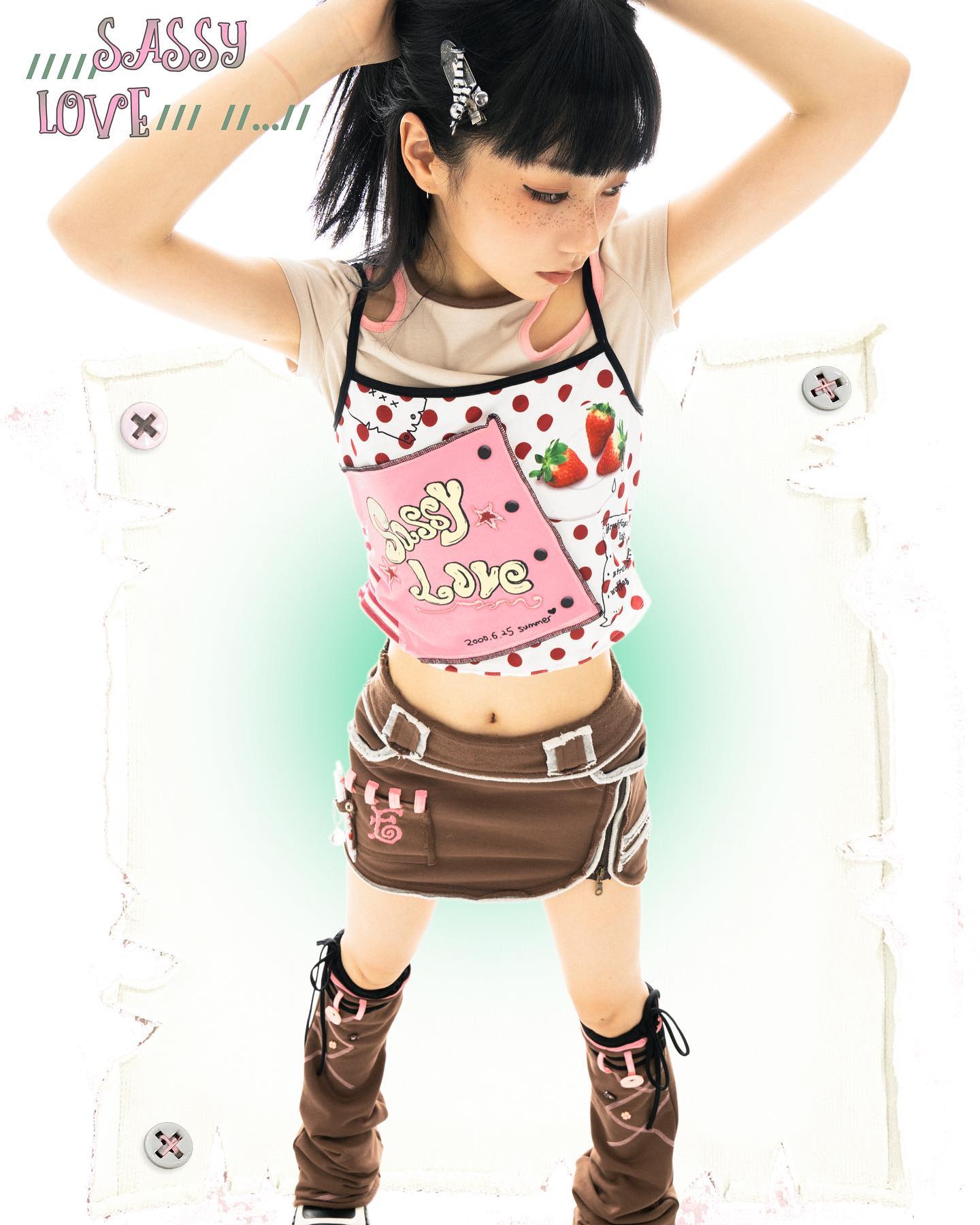 a young girl is modeling a cute and stylish outfit.  she's wearing a sleeveless polka dot top with a pink patch that says "sassy love", brown mini skirt with details, and brown leg warmers.  her hair is styled with hair clips. the overall aesthetic is kawaii and reminiscent of harajuku fashion. the background is a soft mint green with a distressed white frame. the words "sassy love" appear in a stylized font at the top.