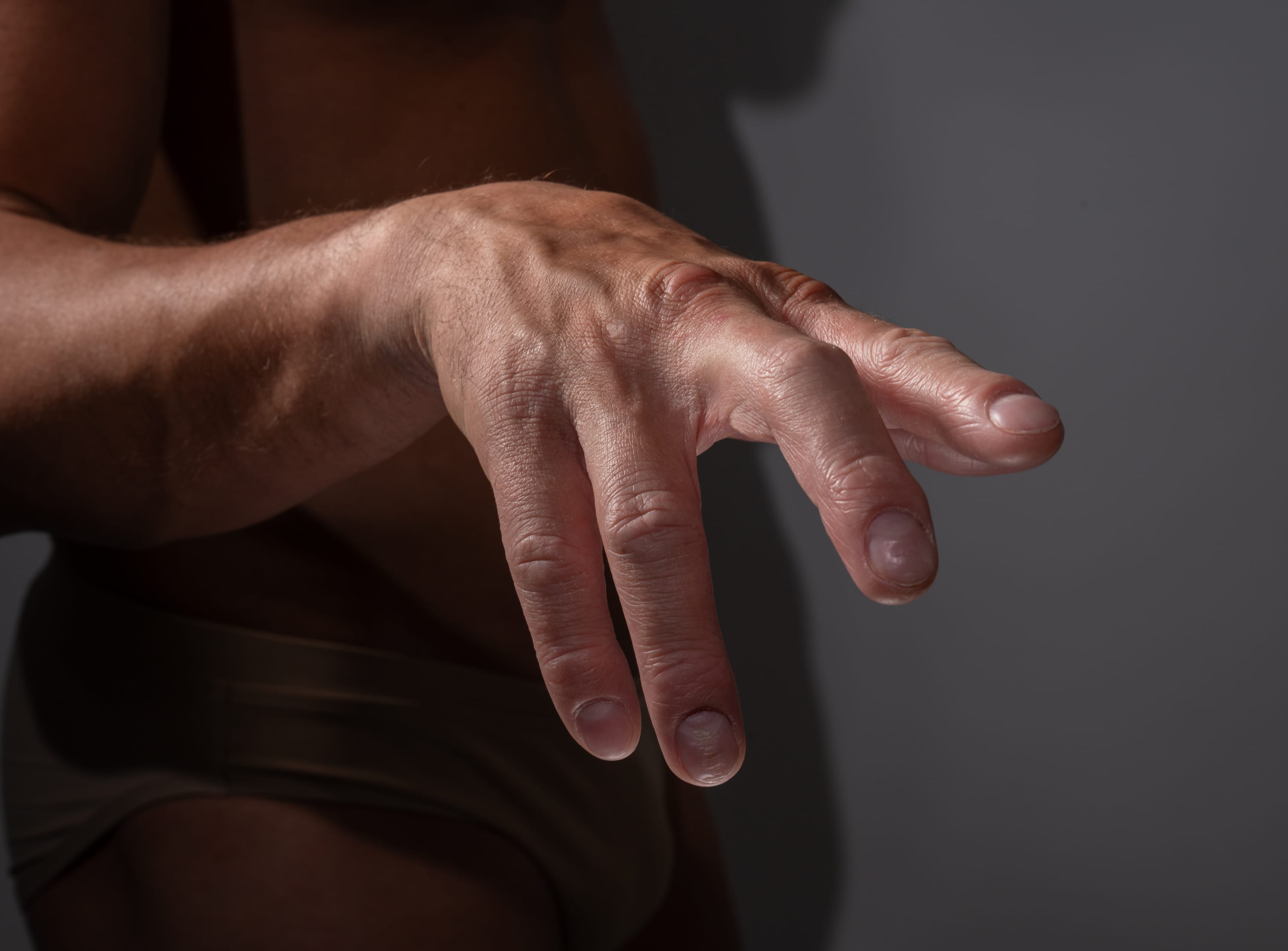 a close up shot of a tanned male hand reaching forward. the image is lit by studio lighting and focused on the man's hand, with the rest of his body slightly blurred out.