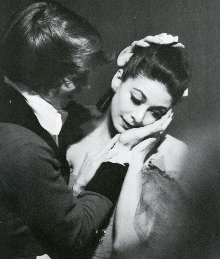 this image is a black and white photograph of a couple, a woman and a man, embracing in a studio setting. the woman, possibly a ballerina, is dressed in a flowing dress and is holding her face in her hands, while the man is