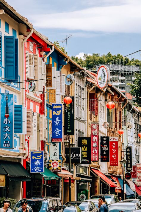 the image shows a vibrant street scene in singapore's chinatown.  rows of colorful shophouses line the street, their facades adorned with various signs in both english and chinese.  red lanterns hang between the buildings, adding to the festive atmosphere. the architecture is a mix of traditional and modern, with some shops displaying contemporary signage alongside older, more traditional elements. the street is bustling, although not overly crowded, with some people visible and cars parked along the roadside. the overall impression is one of cultural richness, history, and vibrant urban life.