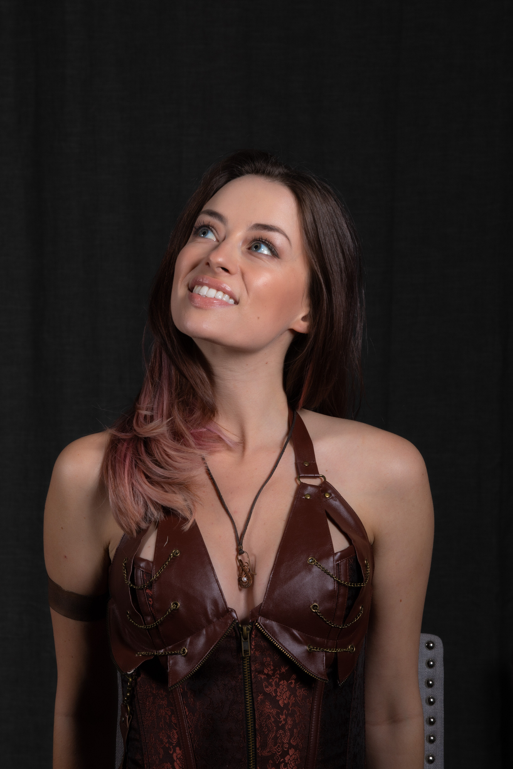 a young woman with pink and brown hair is pictured against a black background. she is wearing a brown leather steampunkstyle corset and a necklace. she is looking upwards and smiling.