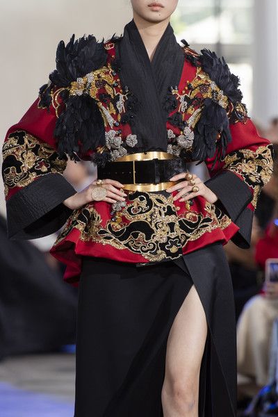 loseup view of a model showcasing a haute couture outfit on a runway. the dress is predominantly red with intricate gold and black embroidery, adorned with black feathers on the shoulders.  a wide gold and black belt cinches the waist. the model also wears gold jewelry. the style is a blend of oriental influences and high fashion. the overall impression is one of elegance, sophistication and intricate detail. the black skirt features a high slit revealing the model's leg. the background is blurred, focusing attention on the outfit.