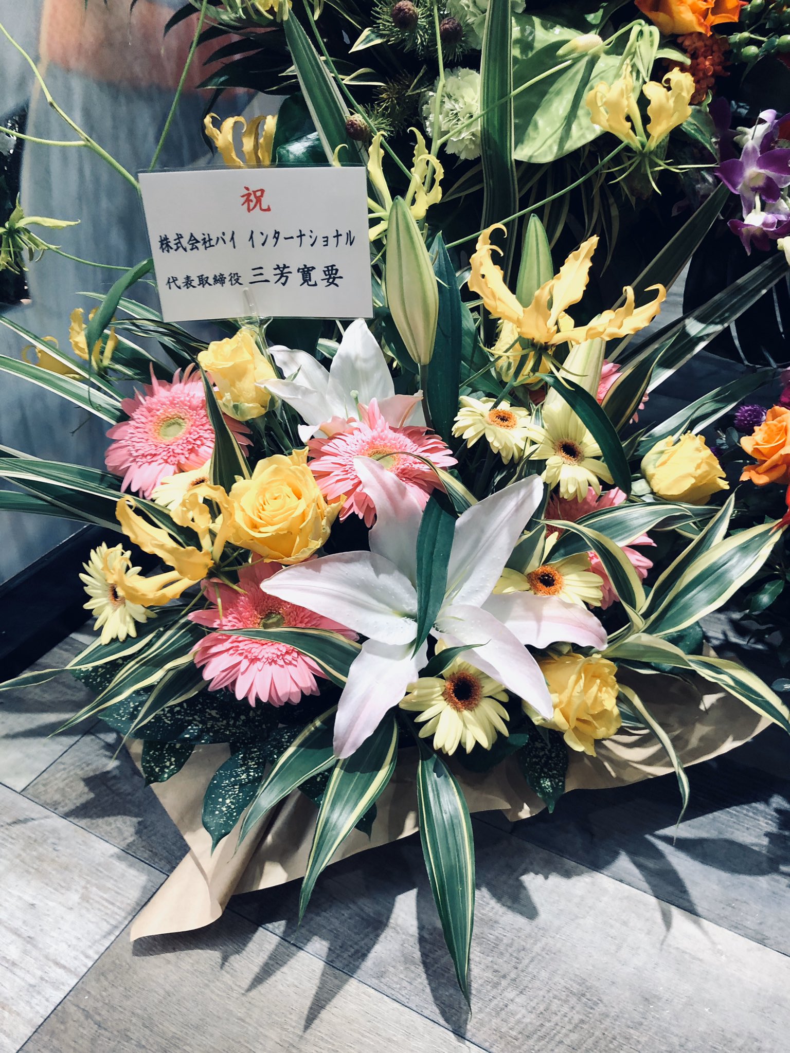 a vibrant flower arrangement with lilies, gerberas, and other flowers is displayed in a congratulatory manner. the arrangement is adorned with long, lush green leaves that extend outward, adding to its visual appeal. a simple white card with japanese text is prominently placed within the flowers, indicating the celebratory nature of the gift. the text on the card suggests it's a congratulatory message for a new opening or a successful endeavor. the setting appears to be indoors, likely a business establishment, as indicated by the tiled floor. the artificial lighting illuminating the scene enhances the vivid colors of the flowers, creating a cheerful and celebratory ambiance.