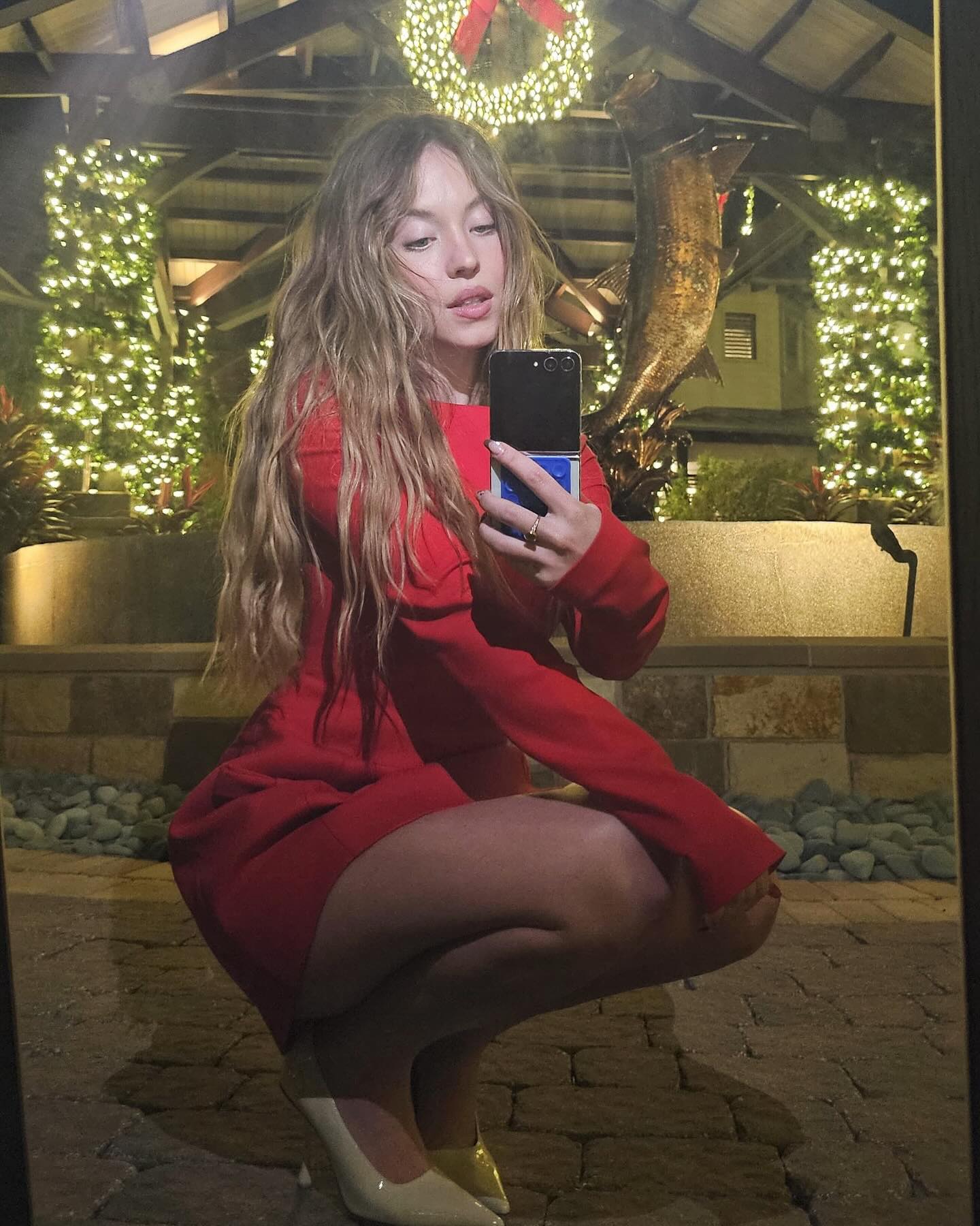 a young woman with long blonde hair is captured in a mirror selfie. she's wearing a short, formfitting red dress and beige high heels. the setting is outdoors at night, with bright christmas lights illuminating the scene, which includes a christmas wreath, and decorative fish sculptures. she is crouching down and holding a google pixel foldable phone, taking the picture. the overall atmosphere is festive and elegant.