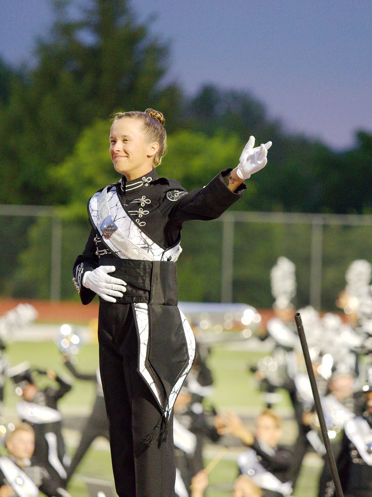 a young woman in a black and white marching band uniform stands in the middle of a marching band field, holding a baton in her left hand, with a slight smile on her face, looking to the right. she is the drum major for the band. the band is performing in a competition outside. there are spectators in the background.