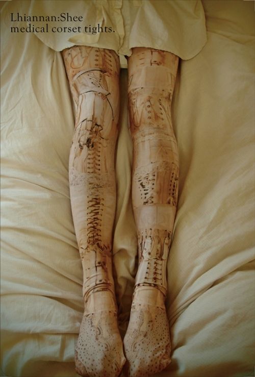 a photograph shows a person's legs clad in lhiannan:shee medical corset tights, lying on a creamcolored fabric, likely a bed. the tights feature an intricate corset pattern printed on them. the tights extend to the feet.