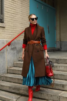 a fashionable woman is wearing a brown coat, red boots, a blue pleated skirt, and sunglasses. she is standing on stairs in an urban setting. she is carrying a handbag in her hand.