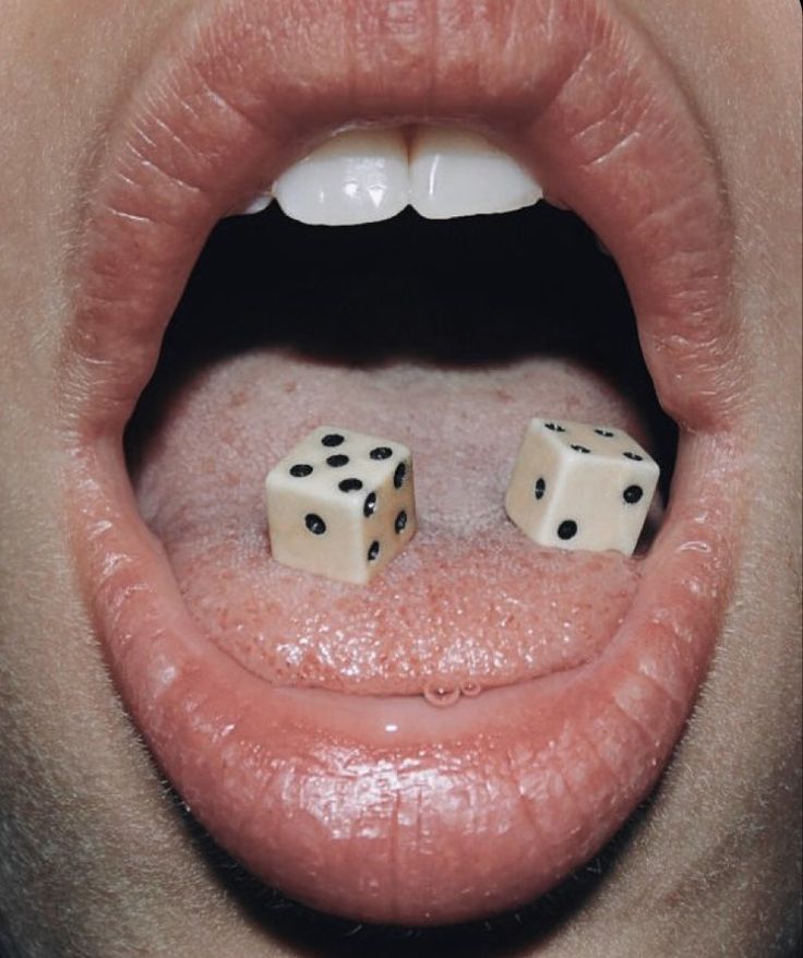 a closeup image of a woman's mouth. her lips are open and she has two dice on her tongue. the image is taken from a low angle.