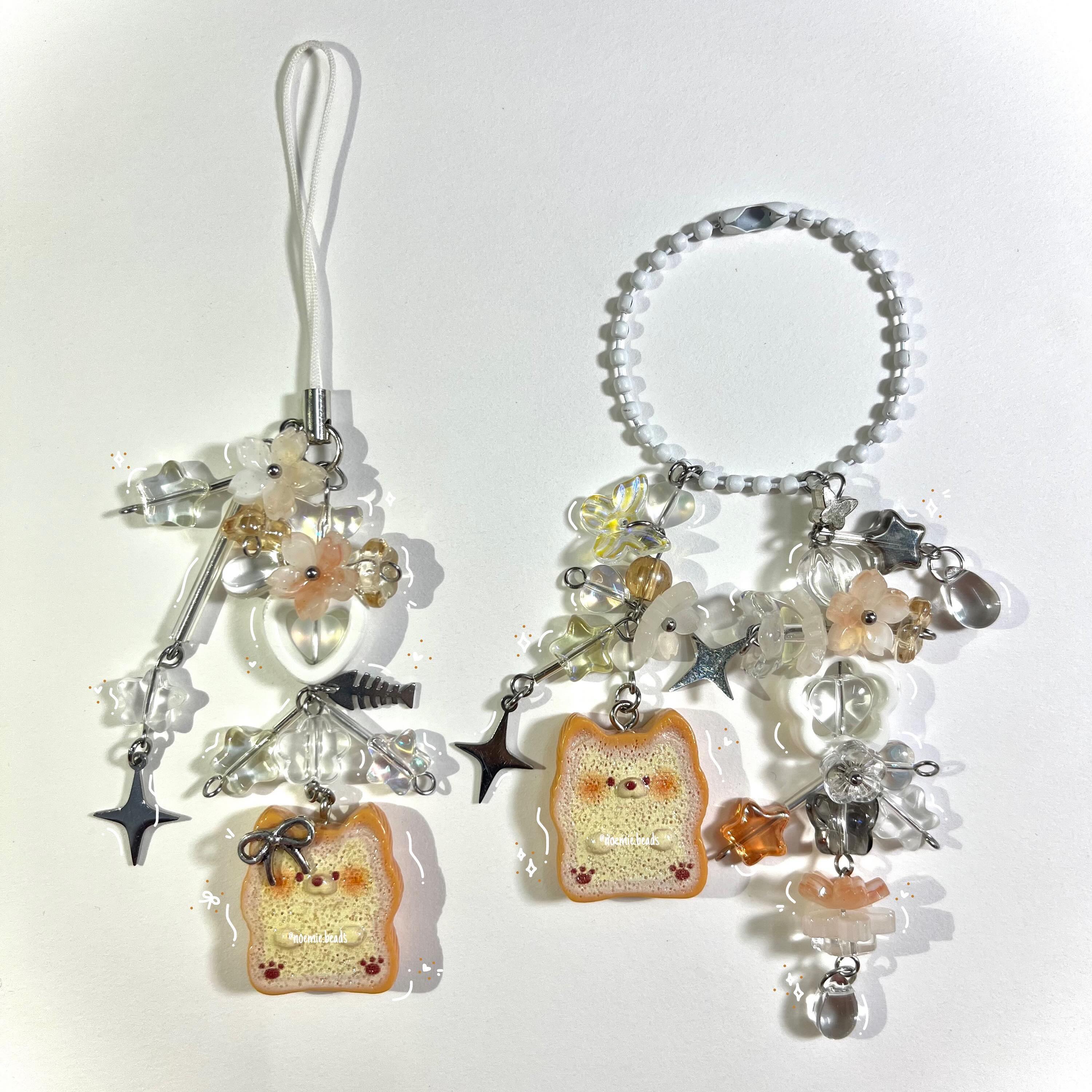 the image shows two handmade phone charms against a plain white background. the charm on the left has a white loop attached to a cluster of clear, gold, and peachcolored flower beads, a silver bar, a white heart bead, a fishbone charm, and a star. at the bottom is a dogshaped pendant resembling a piece of bread with a small bow. the charm on the right has a beaded white loop attached to a similar cluster of flower and clear beads, along with silver star charms and clear teardrop beads. the same dogshaped pendant is at the bottom. the beads and charms create a whimsical and decorative look.
