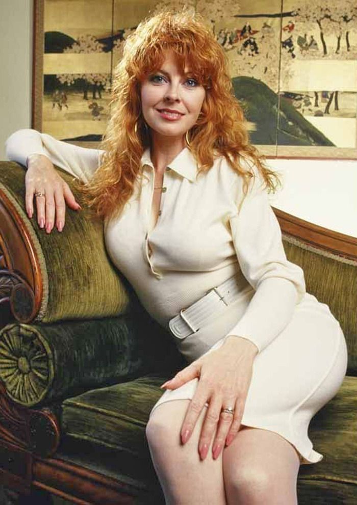 a woman with red hair sits on a green velvet couch. she is wearing a white shirt, white skirt, and white belt. she is smiling.  the background is a wall with a large picture hanging on it.