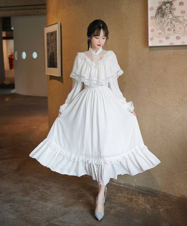 a young woman in a white lace dress is standing in an art gallery. the dress has a high collar, long sleeves, and a flowing skirt with multiple layers of ruffles. she is wearing silver high heels and has her head slightly tilted down. the gallery has white walls and a concrete floor, with artwork visible in the background. the lighting is artificial and bright, illuminating the woman and her dress.