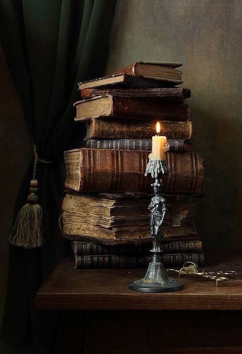 this image features a stack of old, antique books placed on a dark wooden table next to a lit candle in an ornate, antique candleholder. the candleholder has a figure of a person on it. a pair of small scissors are on the table to the right of the candleholder. a dark green curtain hangs in the background to the left, with a tassel dangling from it. the overall ambiance of the image is dark, vintage and moody.
