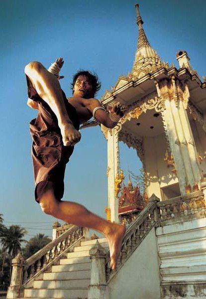 a man wearing traditional muay thai shorts jumps, kicks, and flies through the air, showcasing his mastery of the martial art.  his  action is set against the backdrop of a beautiful white thai temple with intricately carved details and a grand staircase leading up to the entrance. the image captures the energy and dynamism of muay thai, while also highlighting the cultural significance of the sport in thailand.