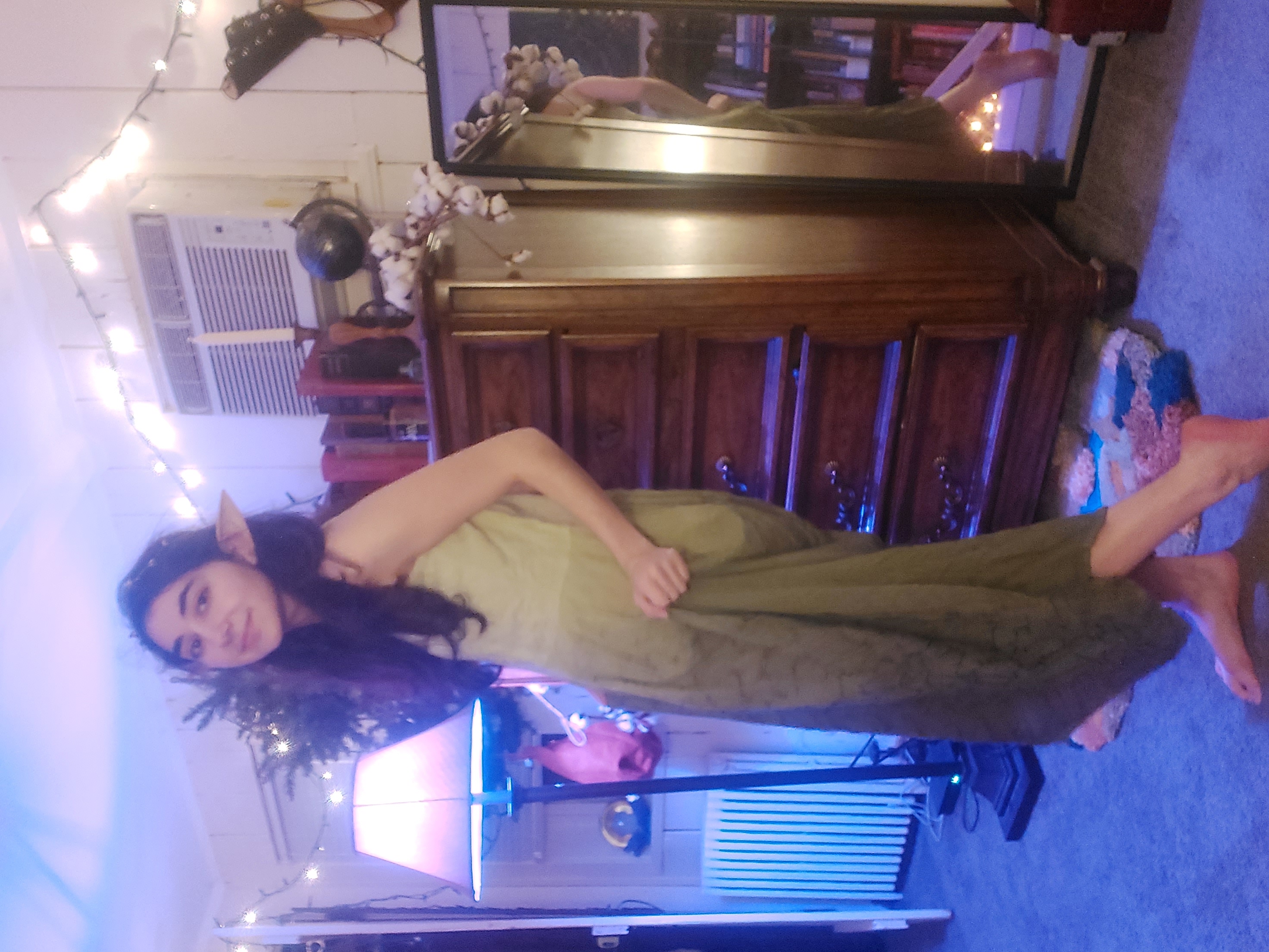 a young woman, dressed in a long, olivegreen dress and wearing elf ears, poses in her bedroom. the room is decorated with christmas lights, and there is a dresser and a mirror visible. she is barefoot and seems to be playfully posing. the overall atmosphere is whimsical and fantasythemed.