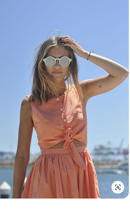 a fairskinned woman with highlighted blonde hair is standing outside on a sunny day by the water.  she is wearing a peachcolored tiefront, buttondown dress with a cutout just above the natural waist.  the dress has a full skirt.  the woman has her left hand on her head, just above her round, light green sunglasses with mirrored lenses.  she is wearing a thin, dark bracelet on her left wrist.  the background is slightly out of focus and includes blue sky and water, as well as buildings and boats in the distance.