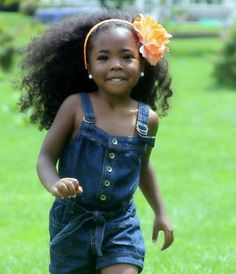 a young black girl smiles at the camera as she runs through a grassy field. she is wearing a denim jumper and a large artificial flower headband. the photo is taken at eye level and is well lit with natural light. the overall tone of the image is happy and carefree.