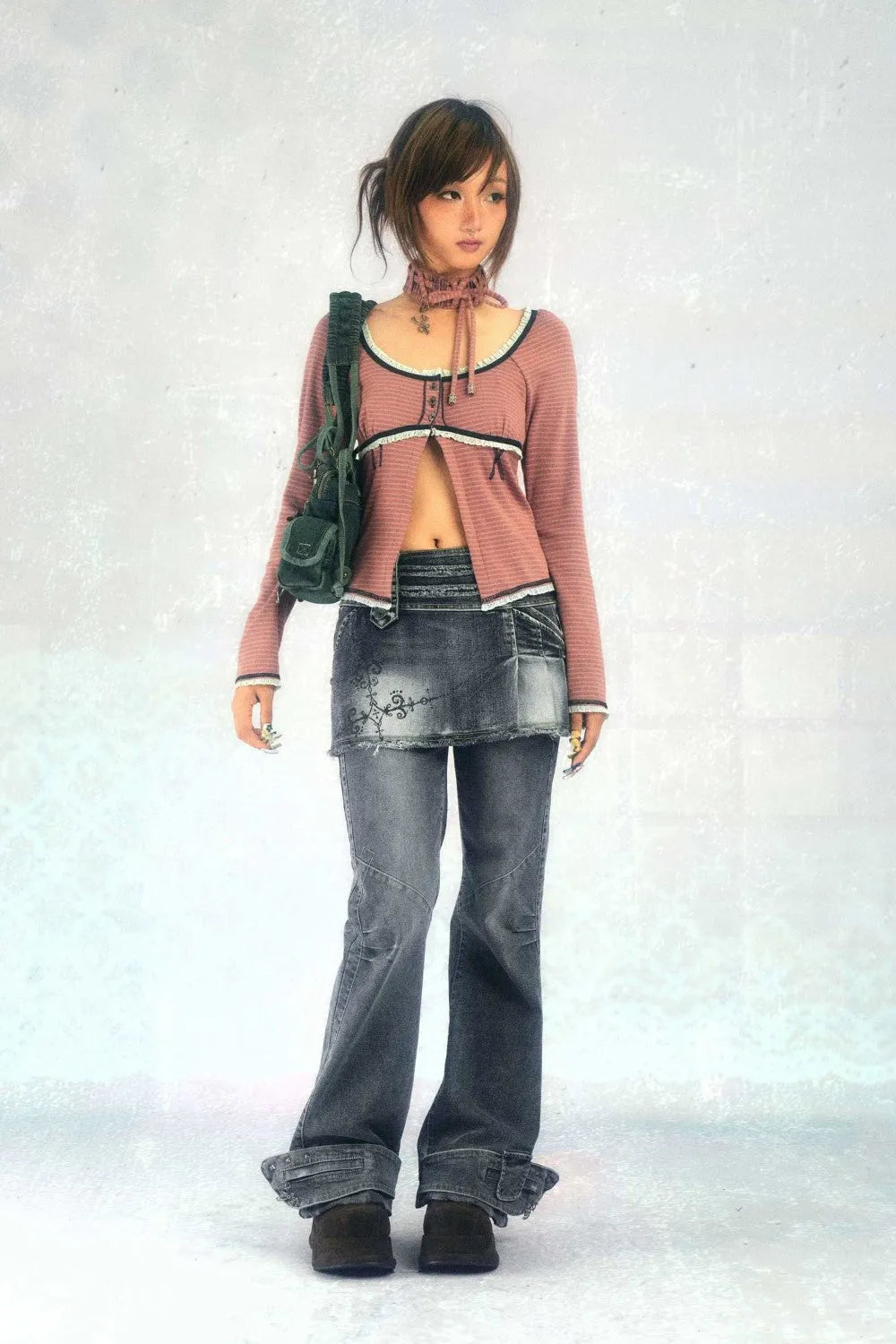 a fullbody shot of a young woman posing against a white background. she is wearing a dusty rose cardigan with a matching denim miniskirt layered over grey bellbottom jeans. her accessories include a dark green shoulder bag, and a crocheted necklace. she has her hair styled in a short, slightly layered bob. her style is reminiscent of early 2000s fashion trends, with a blend of grunge and casual elements.