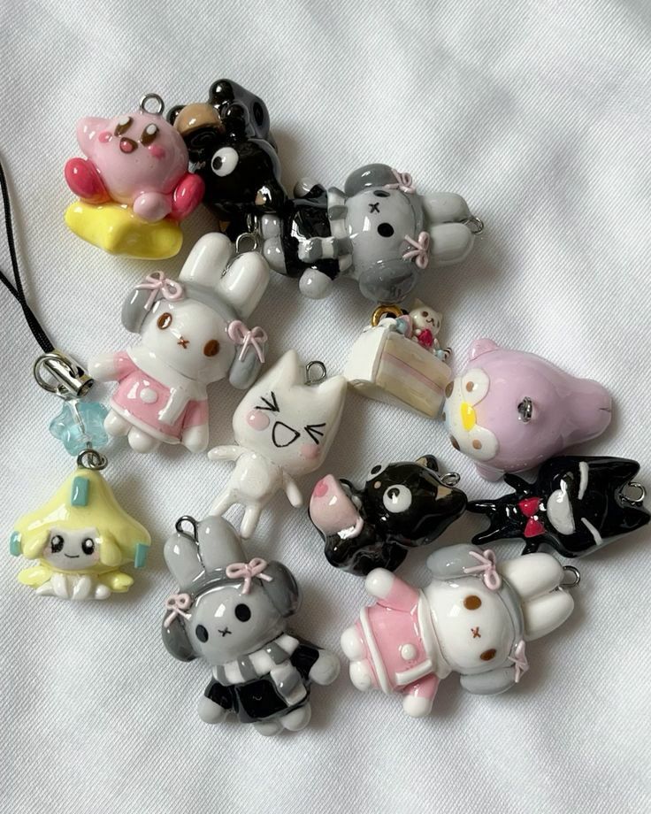 a collection of handmade polymer clay charms are displayed on a white fabric surface. the charms feature various cute cartoon characters, including rabbits, bears, cats, and food items like cake. the colors are predominantly pink, grey, black, white, and yellow. some charms are recognizable as characters like jirachi, kirby, kuromi, and my melody. many are adorned with bows, stars, and other decorative elements. they have loops for attaching to jewelry or accessories.