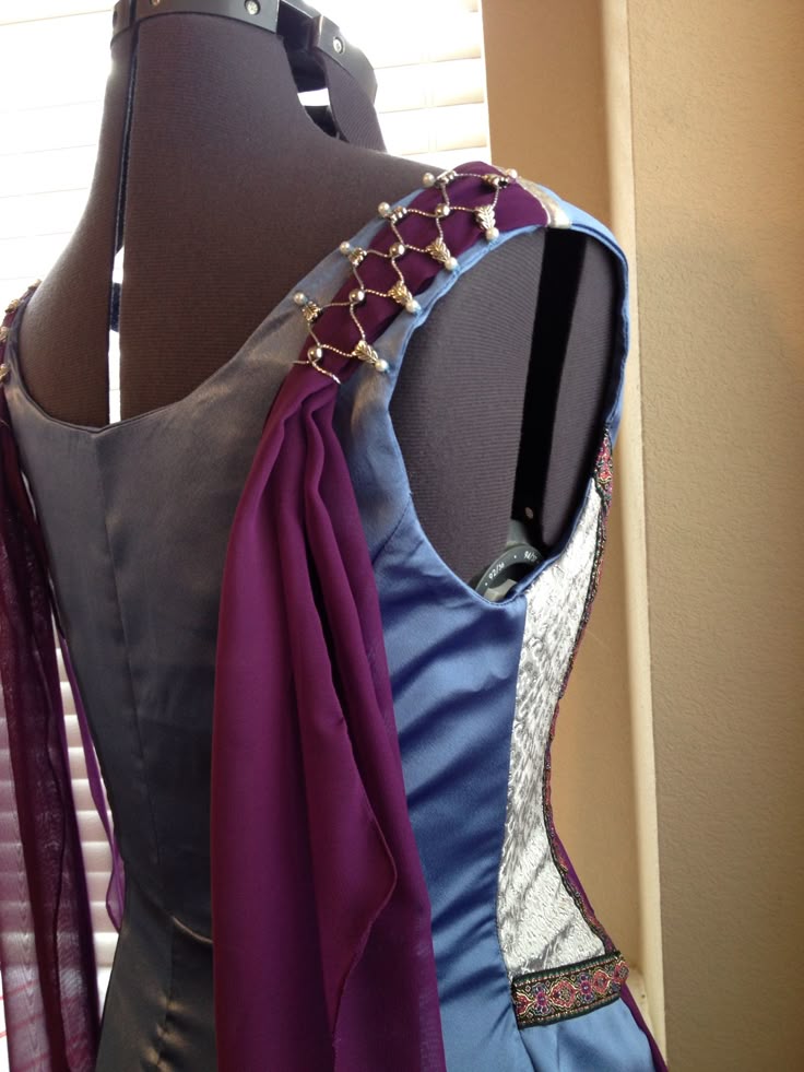 a dress is displayed on a mannequin. the dress is sleeveless and has a blue bodice with silver side panels. the shoulder straps are purple and adorned with beads. a purple sheer fabric drapes from the shoulder.