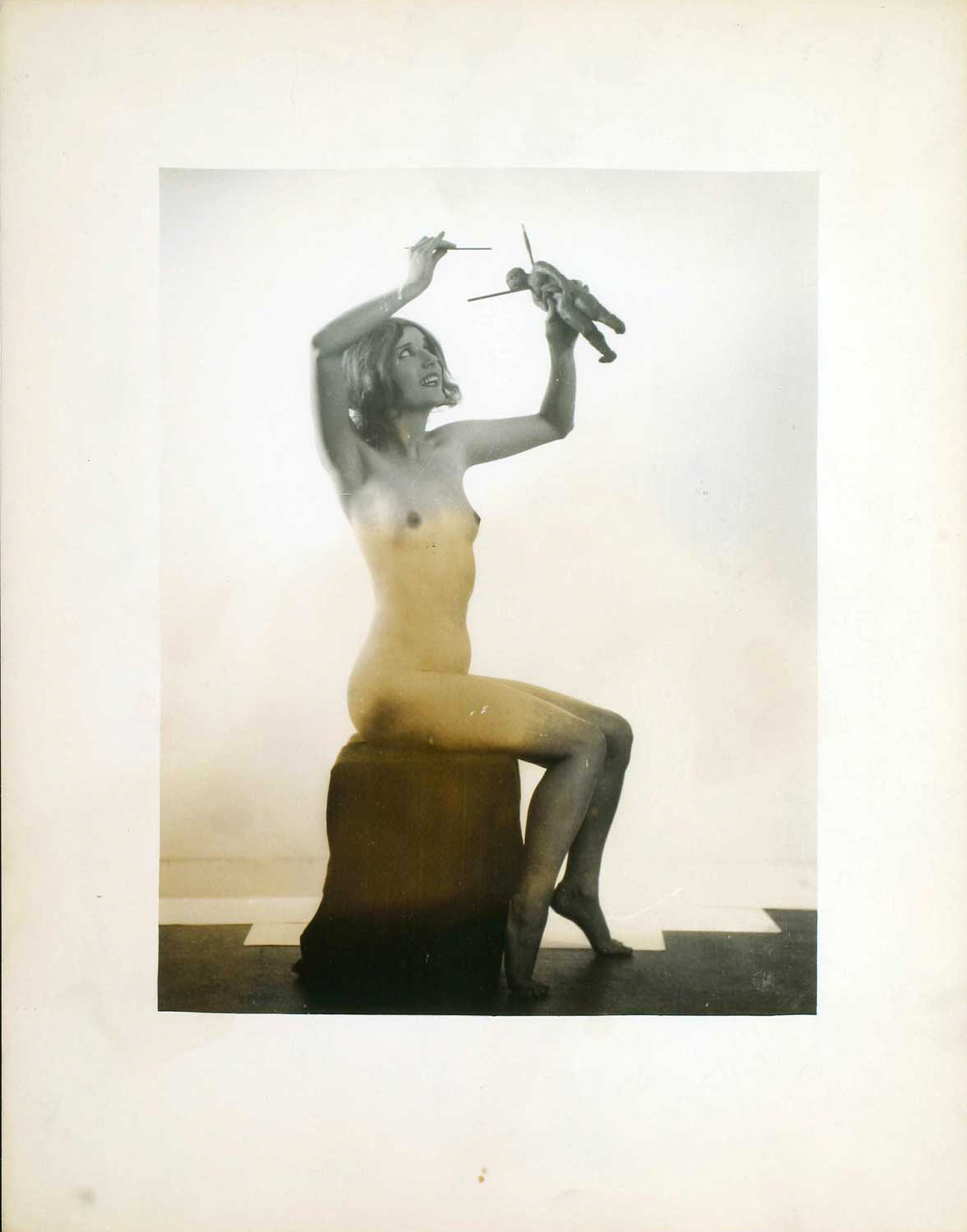 the photograph depicts a nude female model seated on a block. she is holding and manipulating a small toy soldier figure with what appear to be thin sticks or wires above her head. the setting is a minimalist studio space. the image is in black and white/monochrome, with a slightly faded or sepiatoned quality, suggesting a vintage photograph. the overall mood is artistic and slightly surreal. the juxtaposition of the nude female form with the small toy soldier creates a sense of scale and perhaps alludes to themes of art, manipulation, or power dynamics.