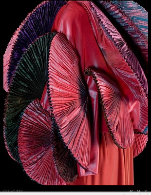 a closeup shot of a red dress with intricate pleated ruffles and a delicate floral pattern. the dress is made of a luxurious fabric and has a very elegant and ornate style. the pleats are gathered in a fanlike pattern, adding to the dramatic and visually appealing nature of the garment.