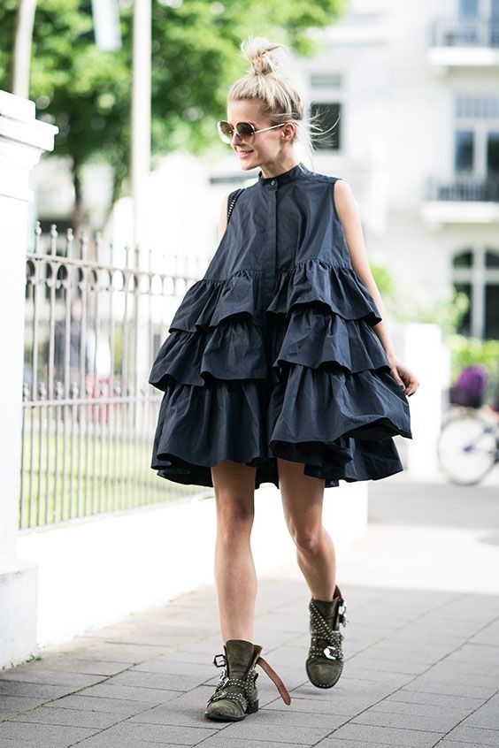 a woman is walking on a sidewalk while wearing a black ruffle mini dress and olive green studded boots. she has her hair up in a bun and is wearing sunglasses.