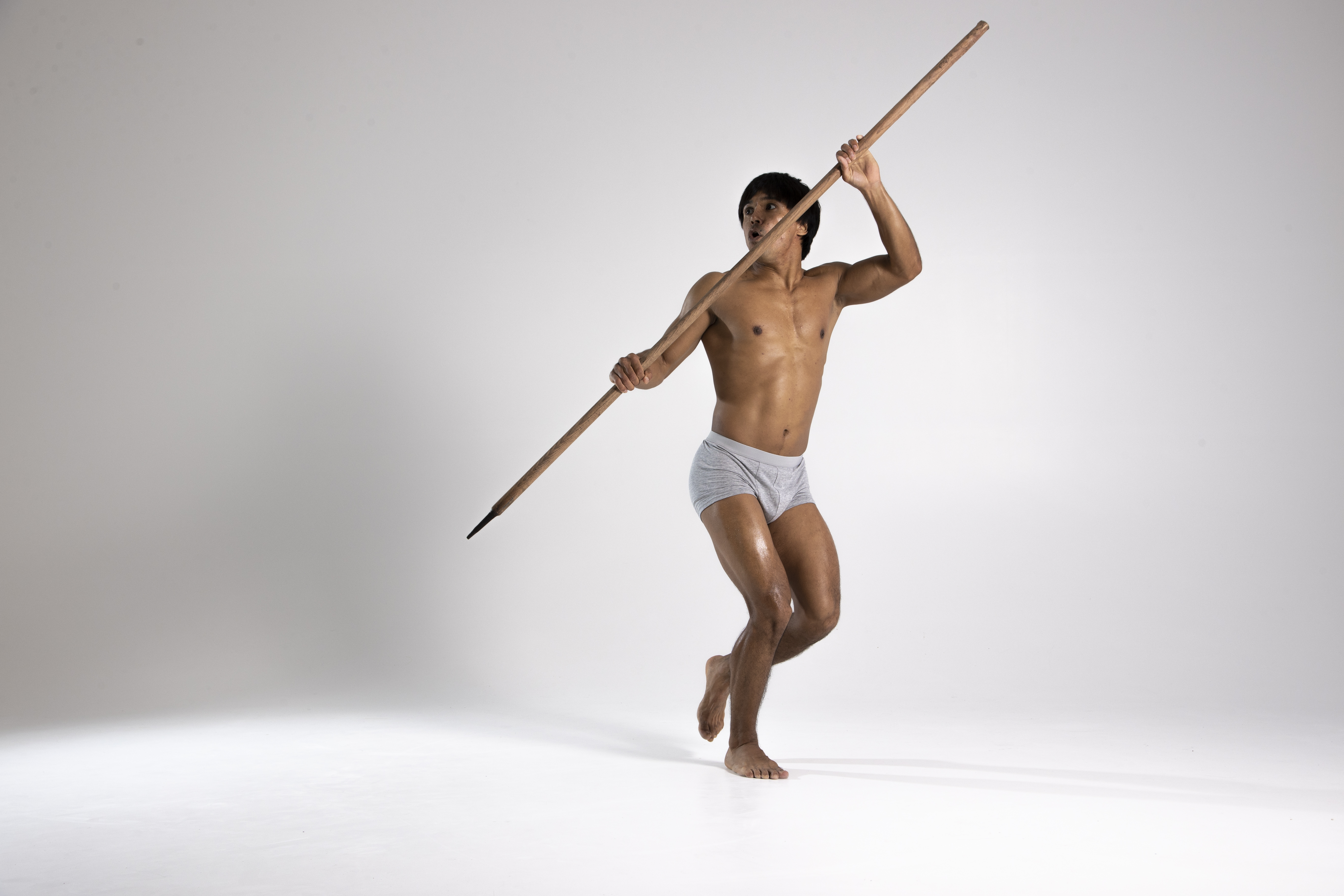 a shirtless athletic man wearing underwear is in motion, holding a spear in a studio shot against a white background. he appears to be throwing the spear, with one foot off the ground.