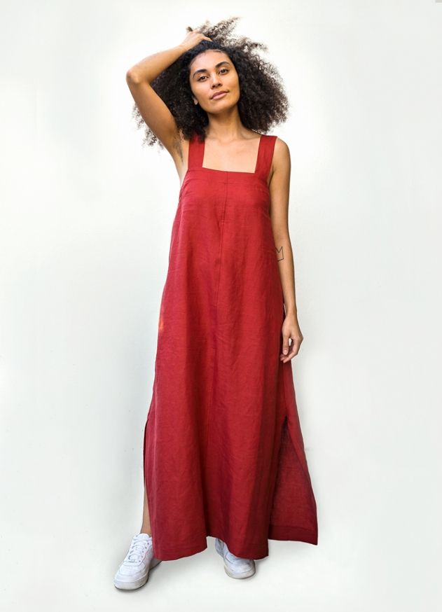 a woman is standing in front of a white background wearing a red maxi dress and sneakers. she has curly hair and is posing with her hand in her hair. the dress has a square neckline and side slits.