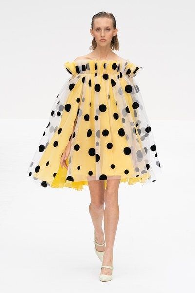 a fashion model walks the runway in a yellow polka dot dress with a tulle cape. the dress has a ruffled neckline and is accessorized with white shoes. the model is wearing her hair down and has a natural makeup look. the background is white and the overall style is very chic and elegant.