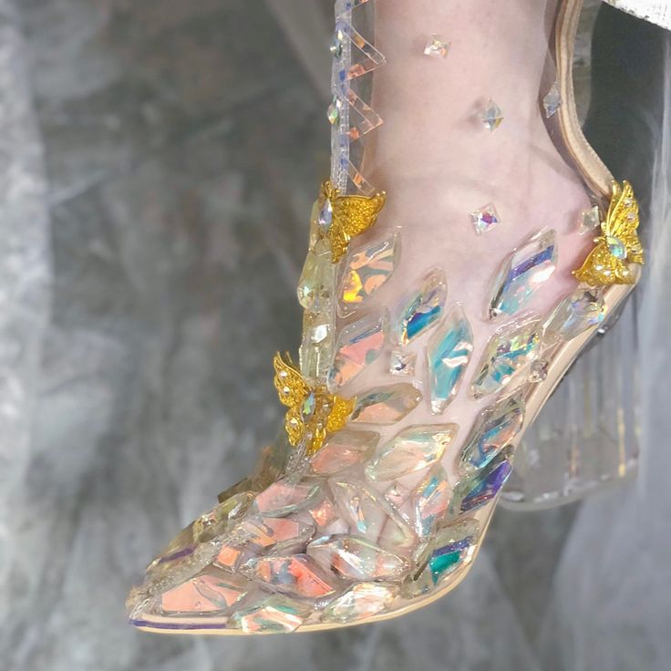 a closeup of a woman's shoe made of glass and crystals. the shoe has a gold butterfly design on the front and is very sparkly and iridescent. it looks very elegant and glamorous.