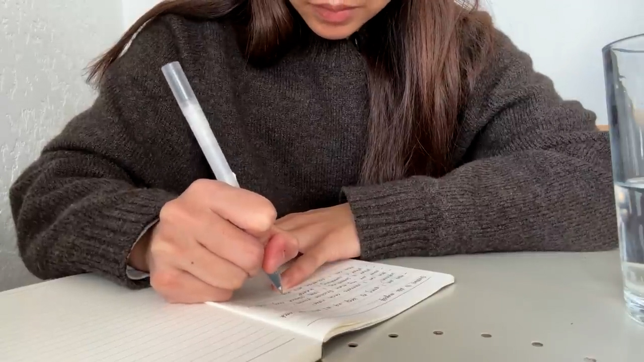 a closeup image of a young woman writing in a notebook.  she is wearing a brown sweater and has long brown hair. the image is taken from a firstperson perspective, so only her hands and the notebook are visible. the lighting is soft and natural, coming from the left side of the image. the overall tone of the image is one of concentration and focus.