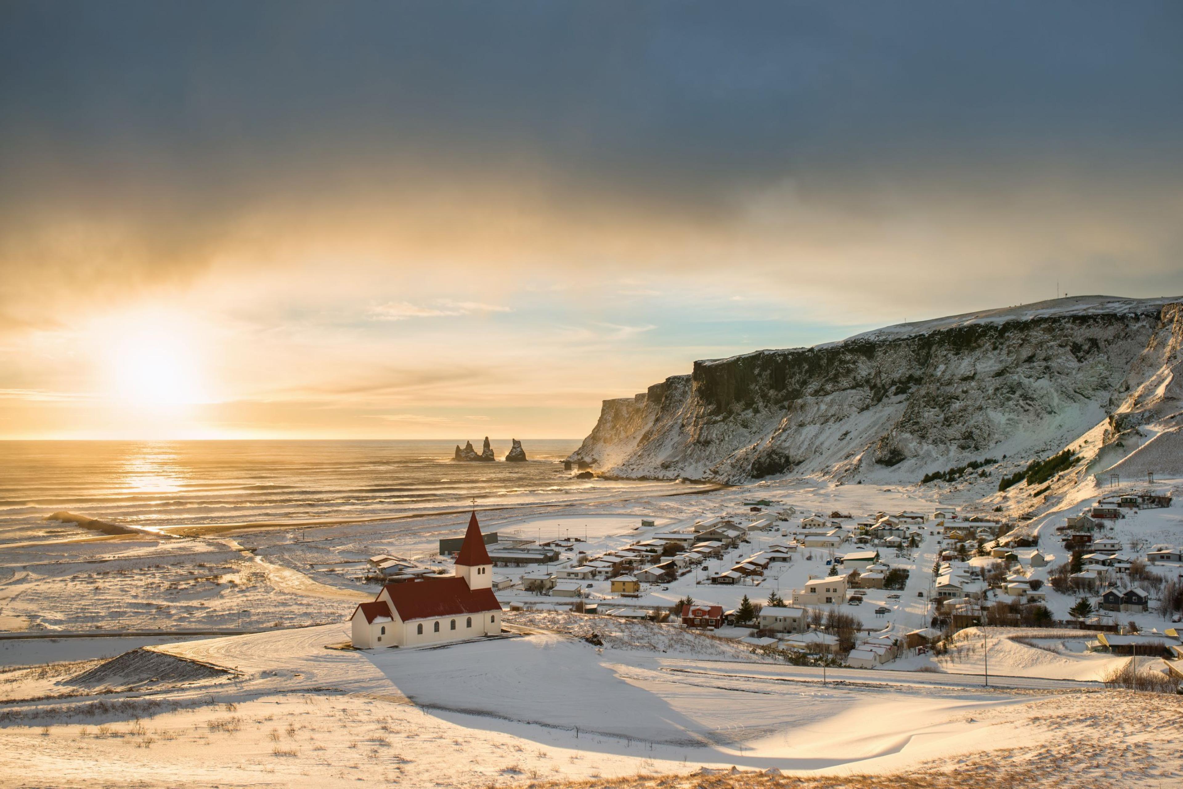 a picturesque scene in iceland with a white church in the foreground, a snowy mountainside, a town nestled below, and a glistening ocean in the distance.  the sunset casts a warm glow over the landscape.
