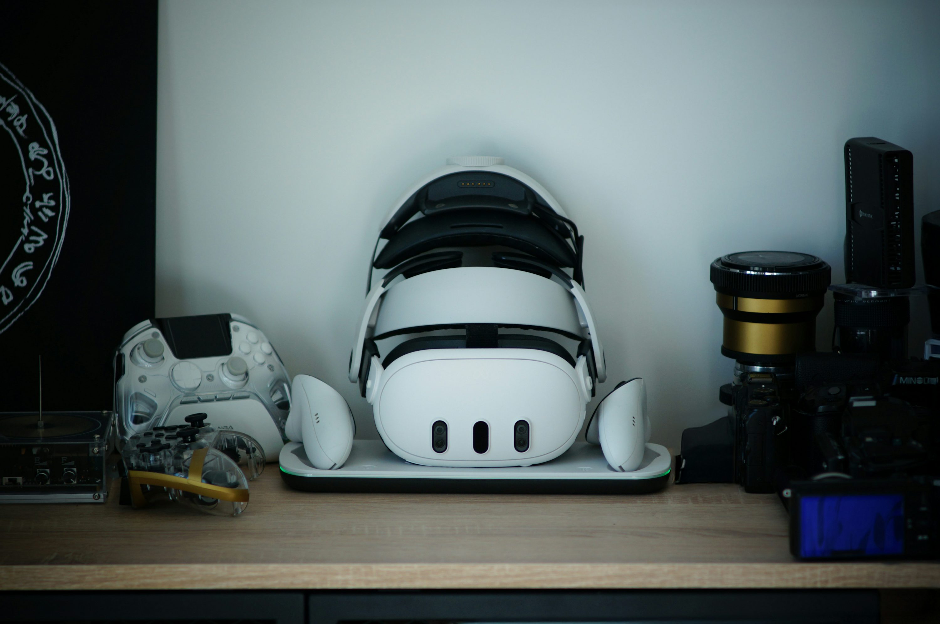 a white virtual reality headset is placed on a charging station in the center of the image. to the left are two gaming controllers and a piece of black and white art. to the right is camera equipment. all are sitting on a light brown desk.