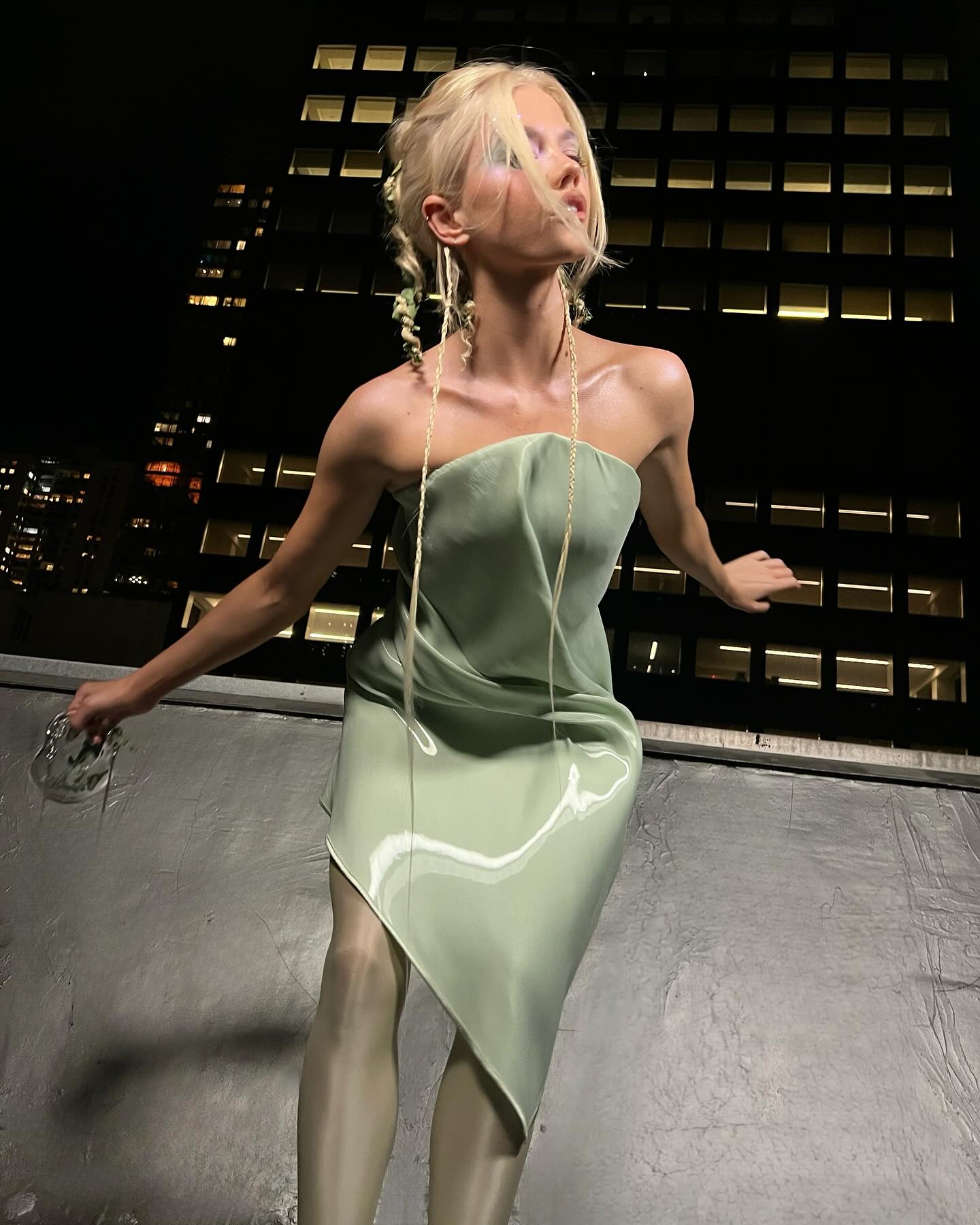 a woman with blonde hair poses in an urban setting at night. she is wearing a strapless, asymmetrical light green dress and shiny tights. she also has long, delicate earrings and is holding a small, clear purse. in the background is a tall building with many illuminated windows.