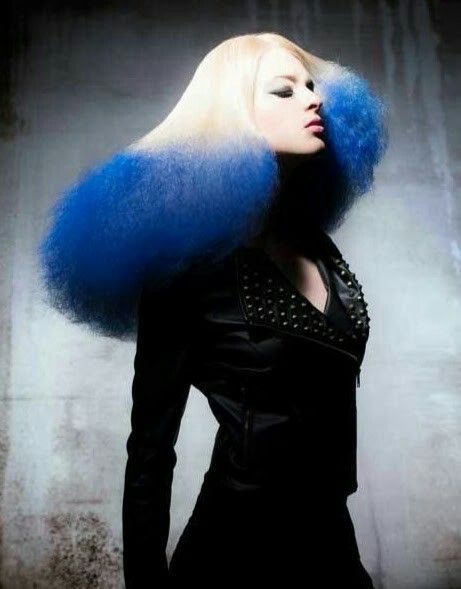 a woman with long blonde hair, dyed blue at the ends, poses in a studio. she's wearing a black leather jacket and a serious expression. the hair style is unique and dramatic.