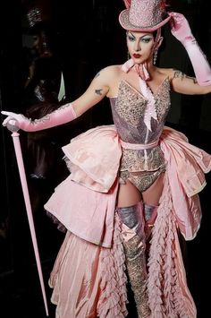 a drag queen in full stage makeup and a pink, elaborately detailed burlesque costume strikes a pose. she is holding a cane in her right hand and adjusting her hat with her left. the background is dark and the lighting is dramatic, highlighting her costume and makeup.