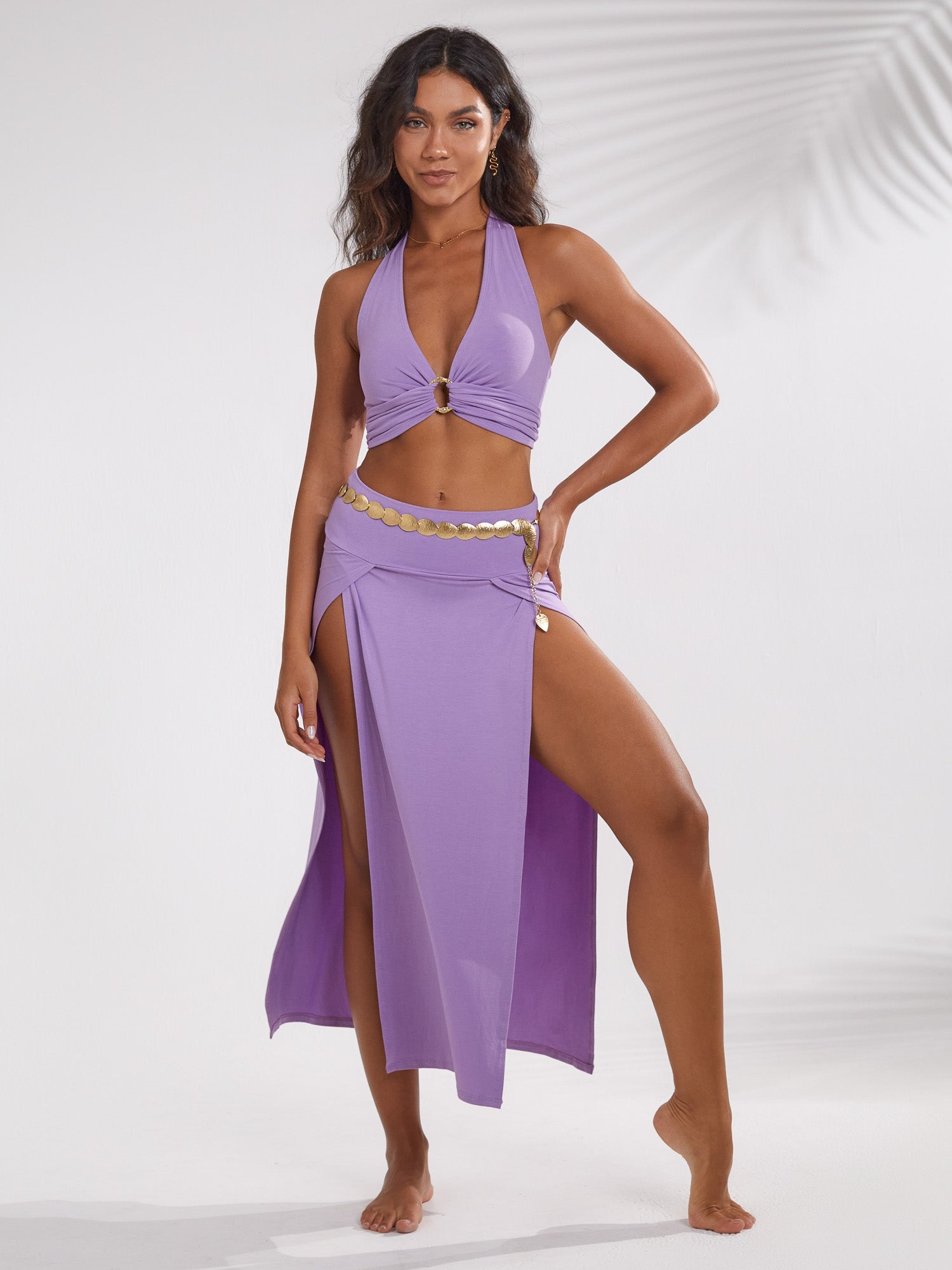 the image shows a woman model wearing a stylish twopiece outfit.  the outfit consists of a light purple halter neck crop top and a matching maxi skirt with high leg slits on both sides. she is also wearing a gold coin belt at her waist. the model is posed against a bright white background, with subtle shadows suggesting soft, diffused lighting. her pose and expression are confident and relaxed. the overall aesthetic is summery, casual, and slightly boho.