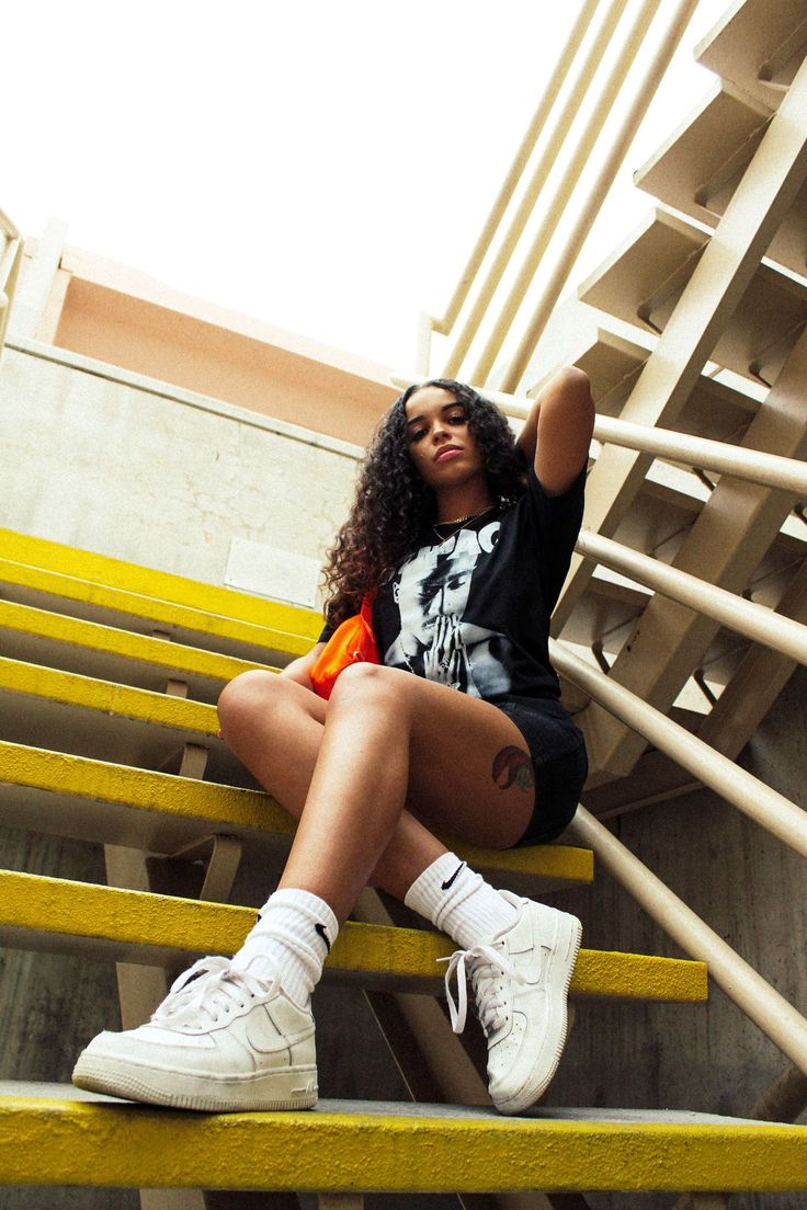 a young woman with curly brown hair sits on bright yellow stairs outdoors. she's wearing a black tshirt with a graphic design, black shorts, white nike air force 1 sneakers, and white socks. she has a small orange crossbody bag and a tattoo on her thigh.  the photo is taken from a low angle, looking up at her. the background features a modern concrete staircase with white railings.  she is looking directly at the camera with a confident expression.